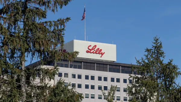 A Lilly sign is seen on the side of a building viewed through pine trees