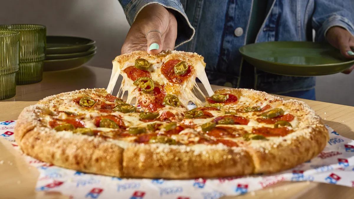 A hand lifts a slice of stuffed crust pizza topped with pepperoni and peppers. The slice trails strings of melted mozzarella.