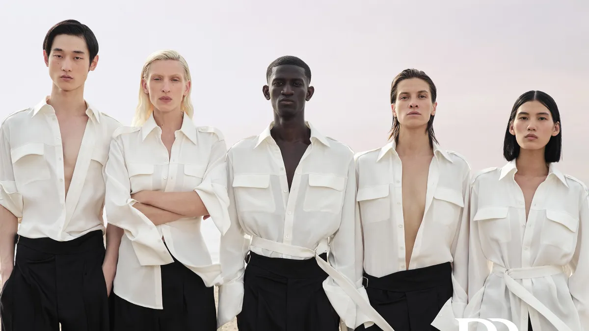 Five models in white shirts and black pants stand on a beach facing the camera.