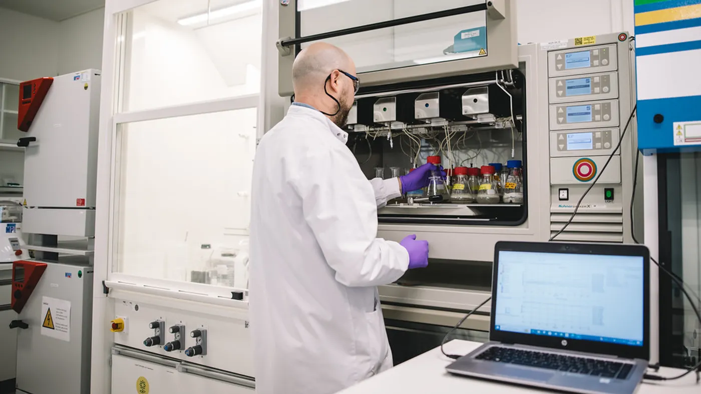 A person in a white coat works in a lab cabinet for packaging research, with a laptop computer to the right
