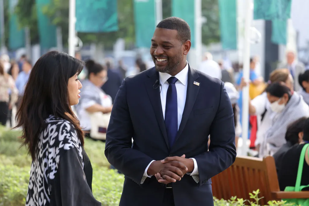 A woman speaks to a man wearing a suit with a U.S. flag pin outdoors, with flags behind them.