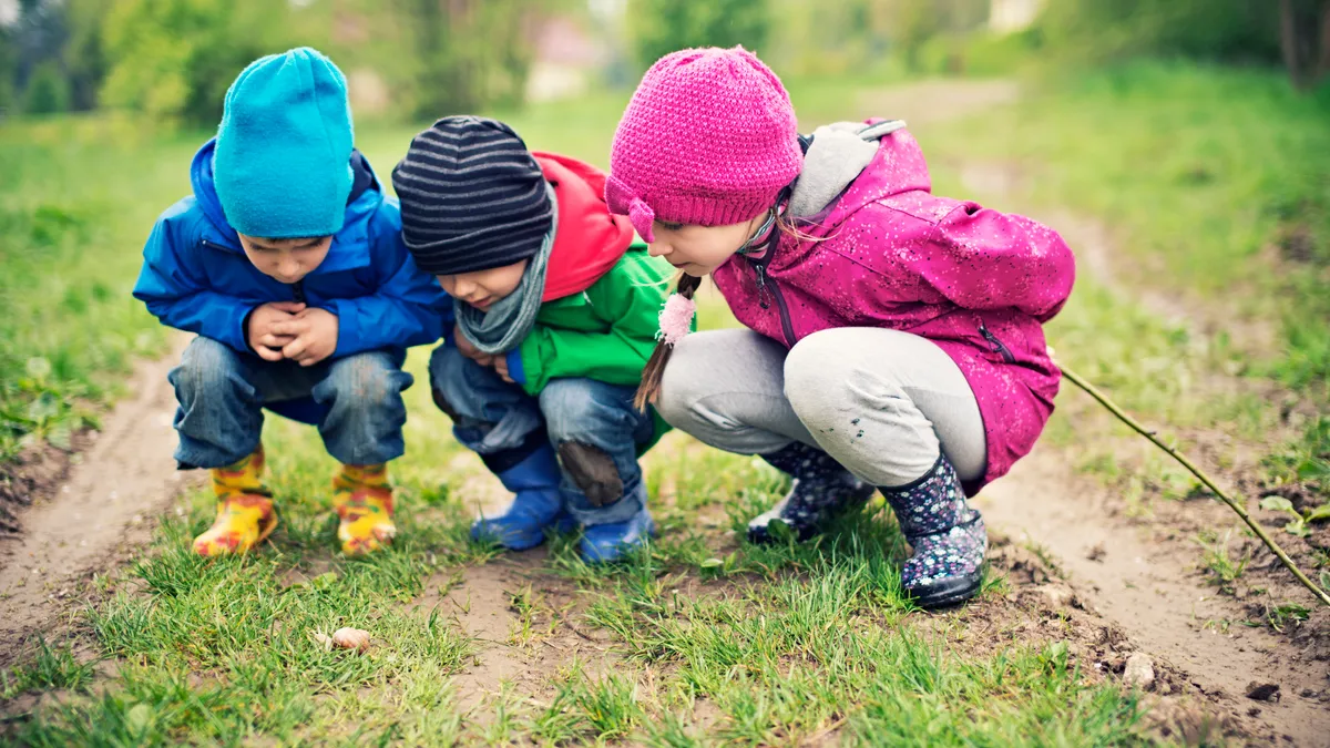 Three young students are outside and leaning close to ground. They are looking down at the grass.