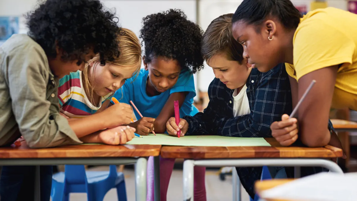 Five students are sitting close together at a table. They have pens and markers in their hands and they are writing on a sheet of paper on the desk.