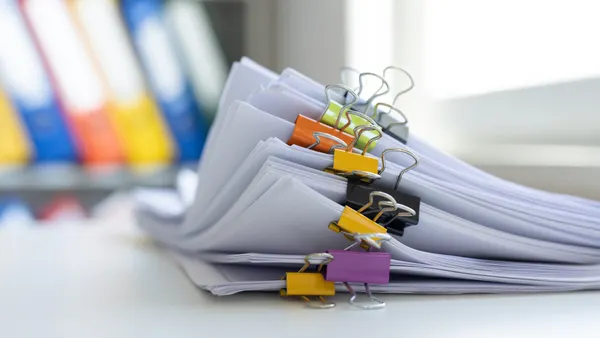 A stack of papers sits on a desk. Several pages are clipped together with colorful clips. A bookshelf with binders can be seen in the background