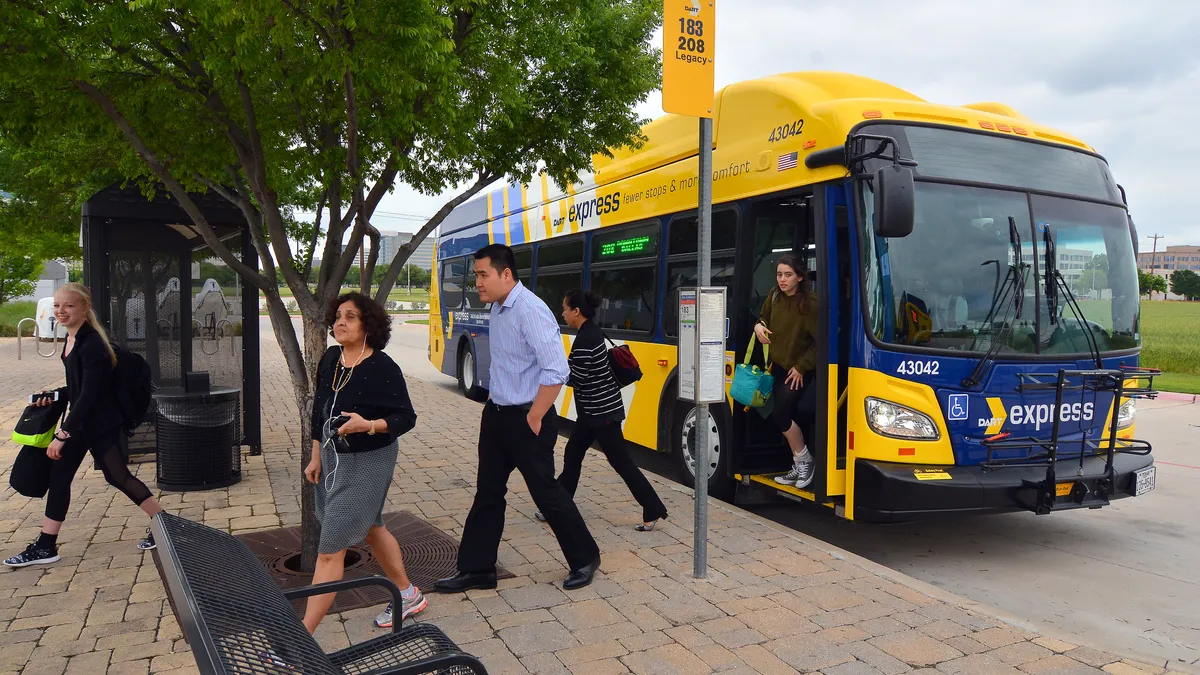 A yellow transit bus marked "express" is at a sidewalk bus stop with five people dressed in light clothes exiting the bus.