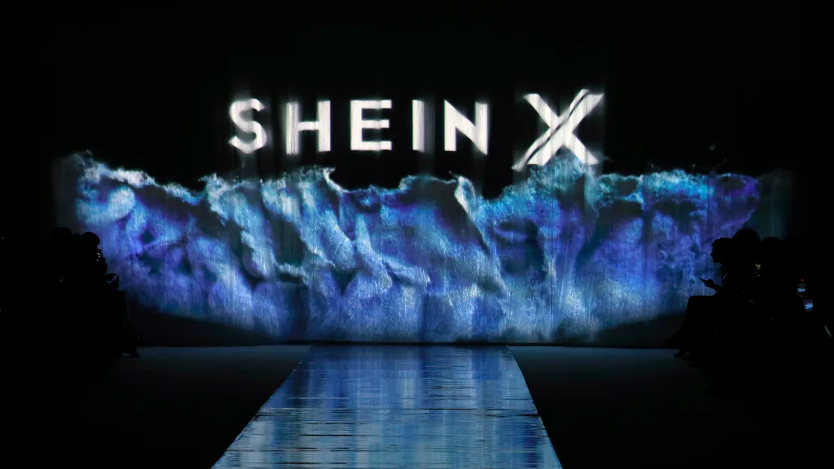 A dark runway is pictured, with no models on it. The words Shein X are displayed and the backdrop is blue, resembling water.