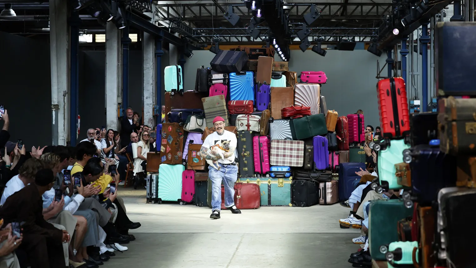 A person stands on a fashion runway holding a small dog. A large pile of suitcases sits in the background.