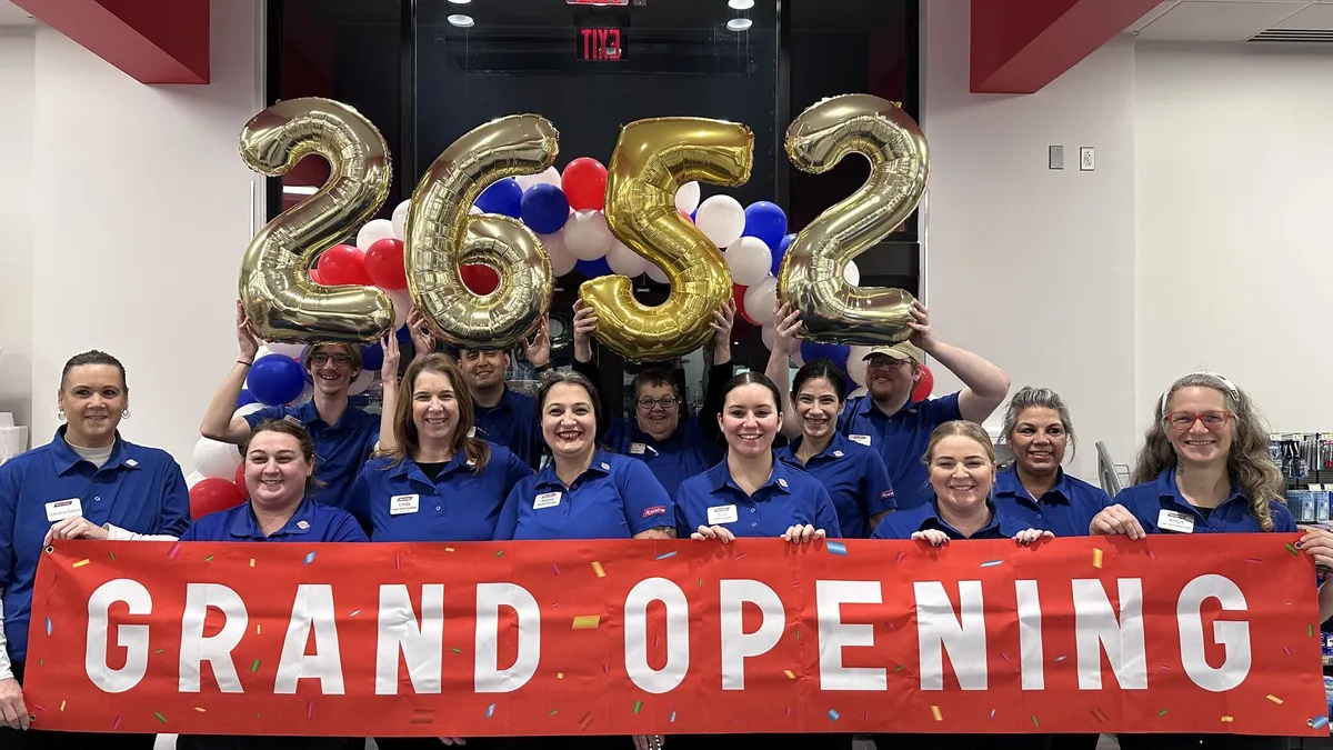 A photo of a number of people in matching blue shirts standing inside a store. Some of them are holding a banner that says "grand opening" while others are holding balloons that say "2652."