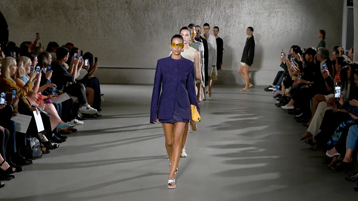 On a gray runway, a model in a navy miniskirt and suit jacket wears sunglasses and carries a yellow clutch as other models follow behind. A crowd watches from the sides.