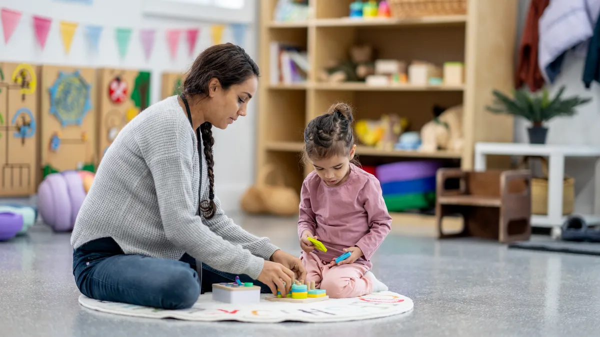 A young student and an adult sit on the floor in a classroom working with colorful blocks