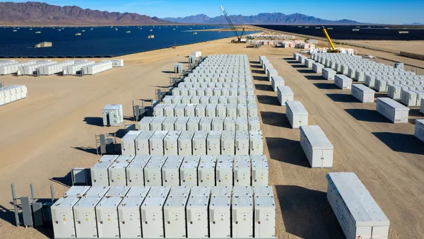 Containers of batteries sit in a desert next to a field of solar panels.