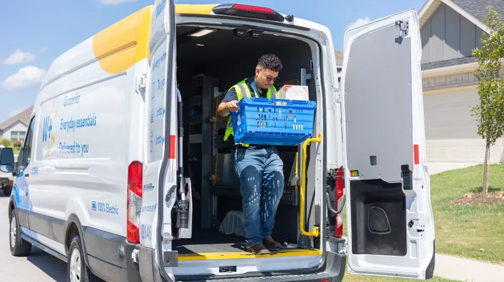 Man holding a blue crate exiting Walmart delivery van from back doors.