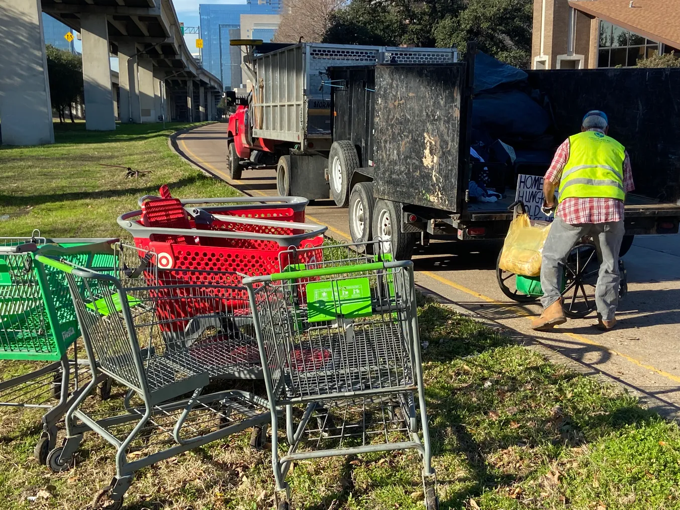 A person in a yellow vest loads items into the pack of a truck parked below a raised freeway. A group of shopping carts sit on the grass nearby.