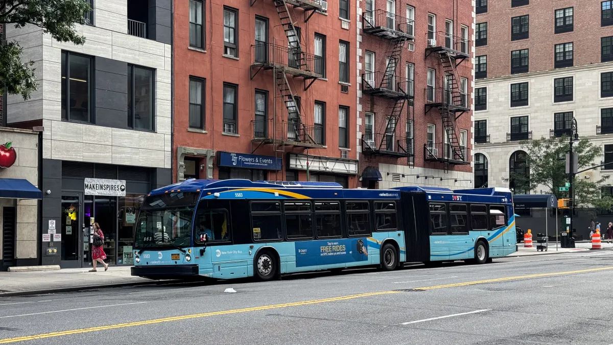 An articulated blue bus stopped at a curb with a woman on the sidewalk and apartment buildings with fire escapes behind.