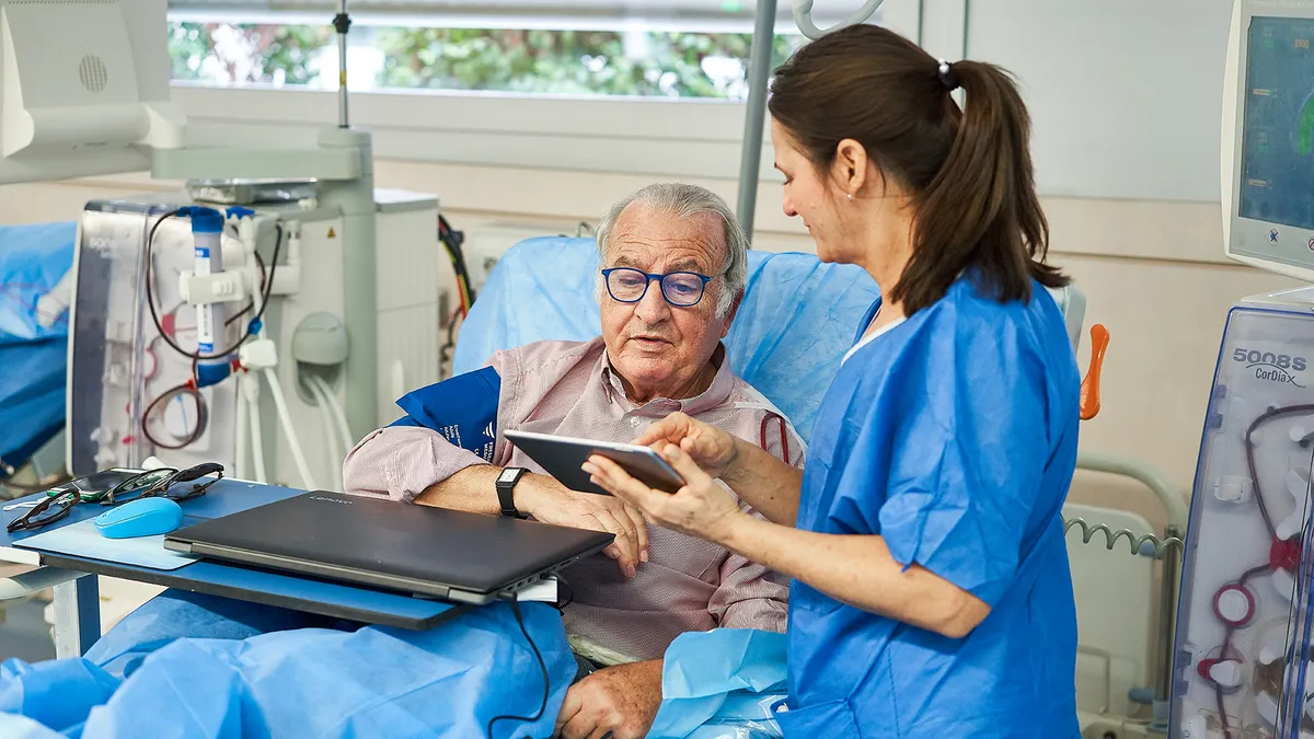 A person wearing blue scrubs holds a tablet, and another person who is sitting looks at it.