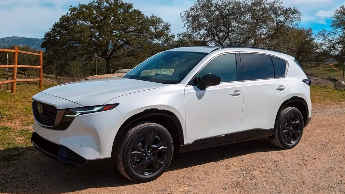 A white 2026 Mazda CX5 crossover parked outdoors with a wooden fence, trees and hills in the background.