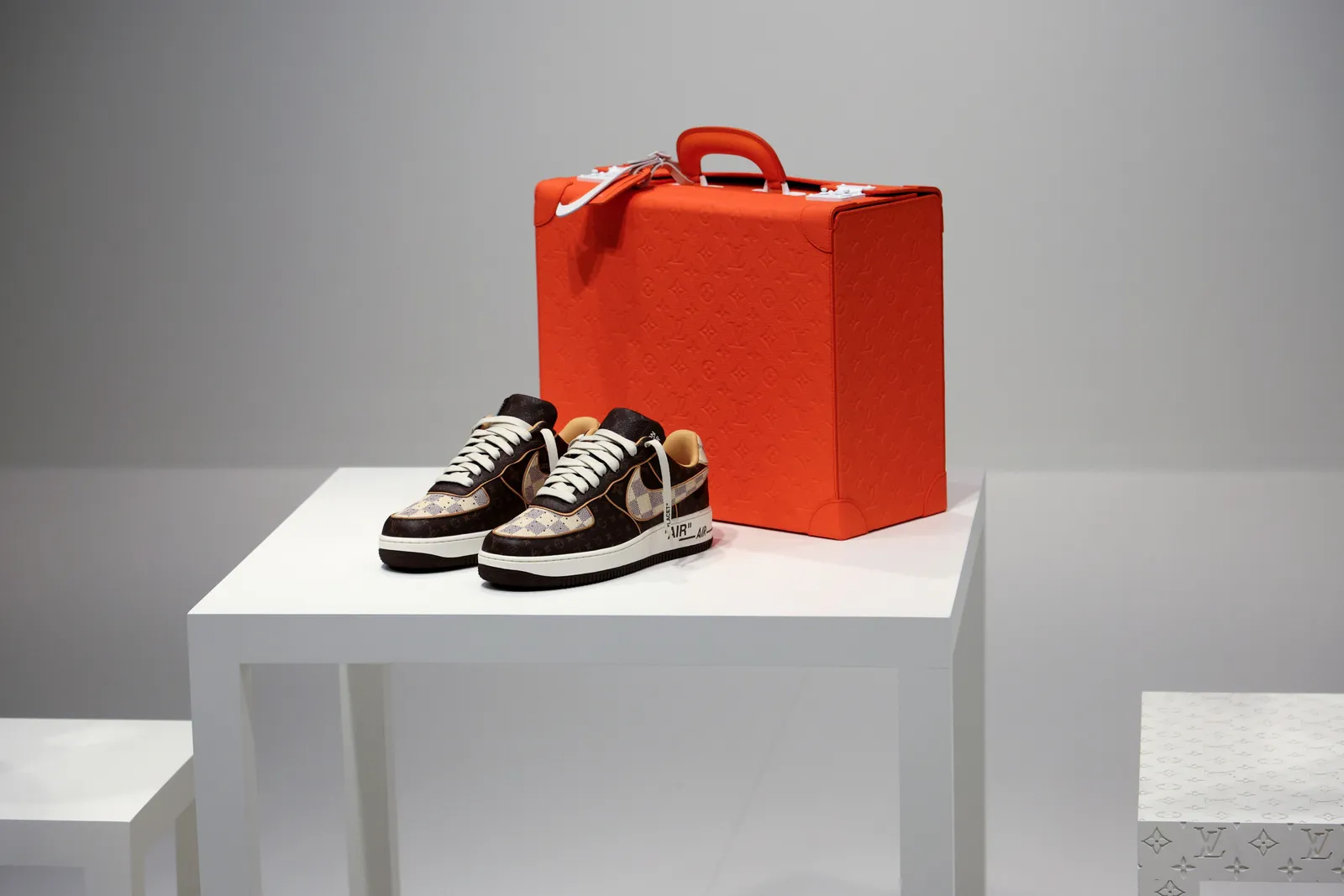 Minimalist display of logo-covered Louis Vuitton & Nike "Air Force 1" sneakers by Virgil Abloh on a gray podium in front of an orange valise.