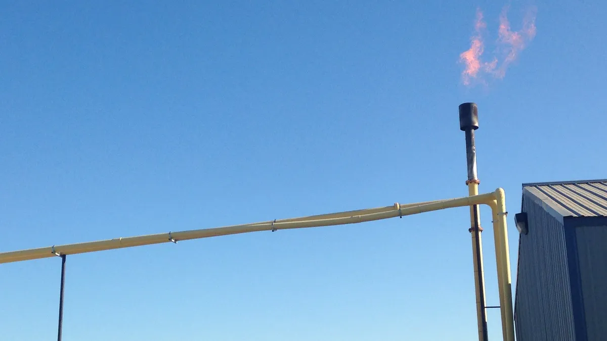 A flare burns gas coming out of a thin metal pipe, part of a system attached to a metal shed.