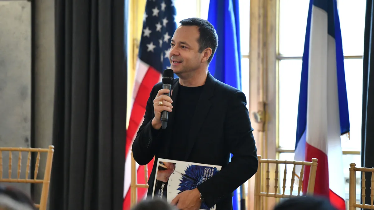 A man in a suit jacket stands in front of several national flags and holds a microphone.