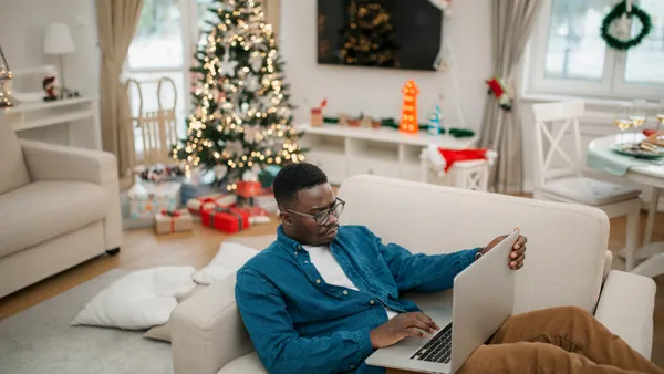 Person sits on an oversized chair with a computer and a Christmas tree, presents and Santa hat in the background.