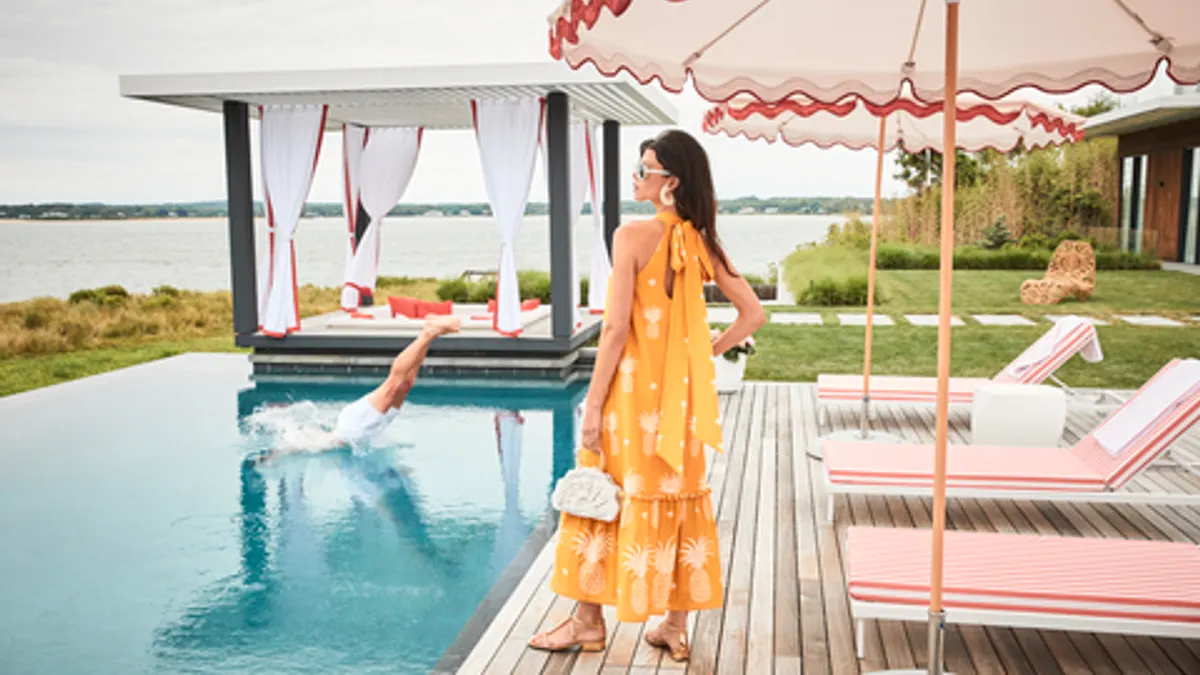 A person stands poolside wearing an orange maxi dress and sunglasses.
