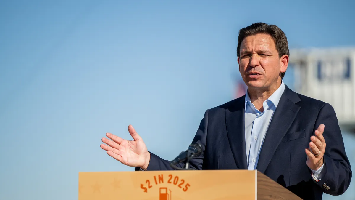 Ron DeSantis speaks in a navy blue suit and light blue shirt, at a wooden podium that reads "$2 in 2025" in red about a red gas can over the words "Fueling American Freedom" in blue.
