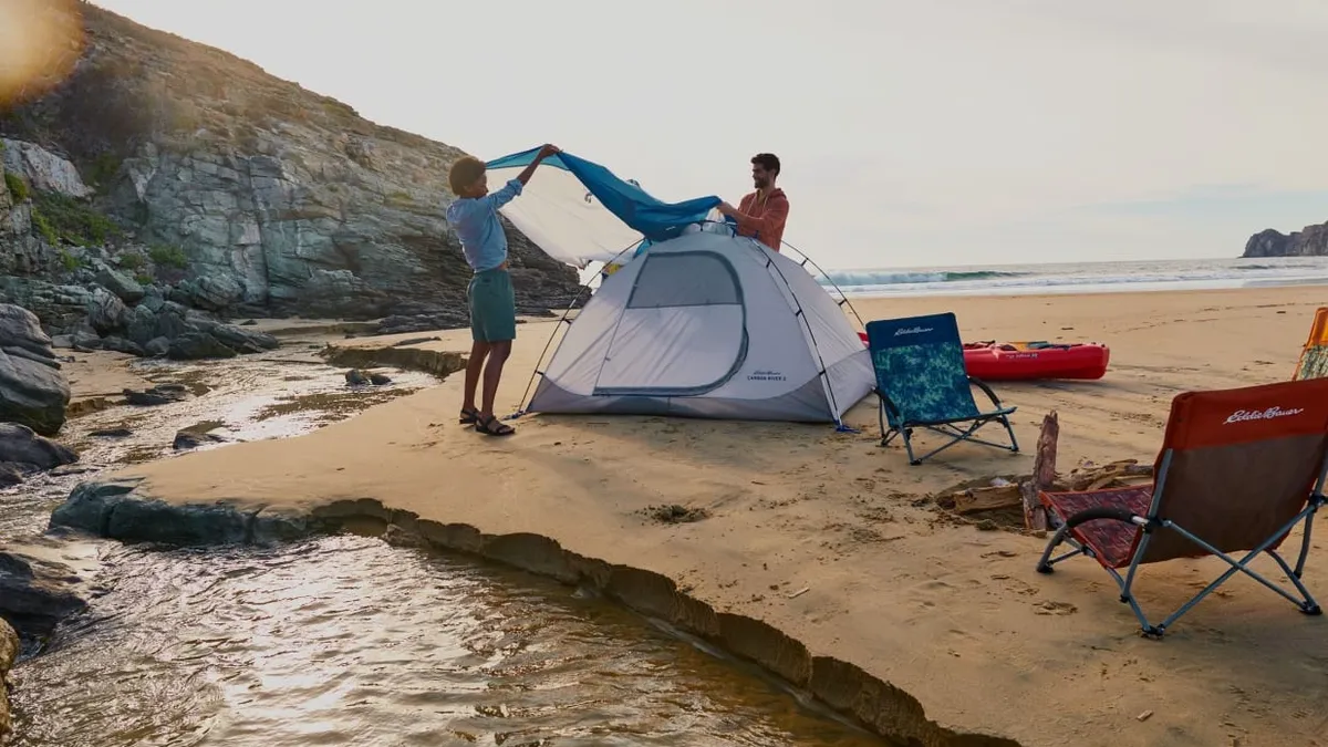 Two people with a tent and folding chairs stand on the beach.