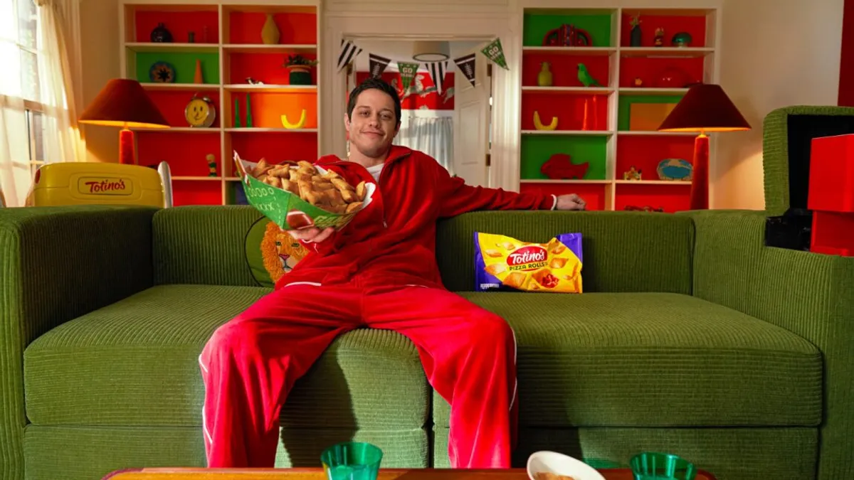 Totino's ad featuring Pete Davidson, who is seen sitting on a green couch holding a bowl full of pizza rolls in a room decked out with Totino's merch.