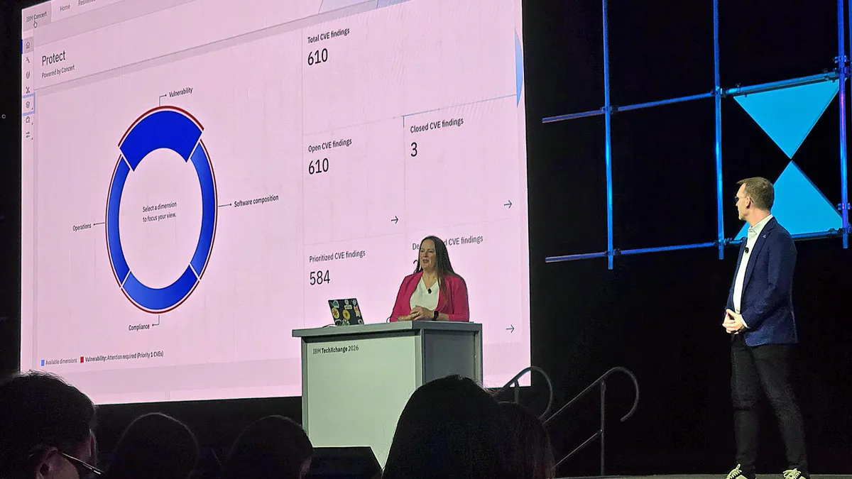 Two people stand on a stage in front of a large screen showing a software UI. One is centered behind a podium wearing a pink jacket. The other is to the right wearing a blue jacket. Some of the audience members' heads are visible in the foreground.