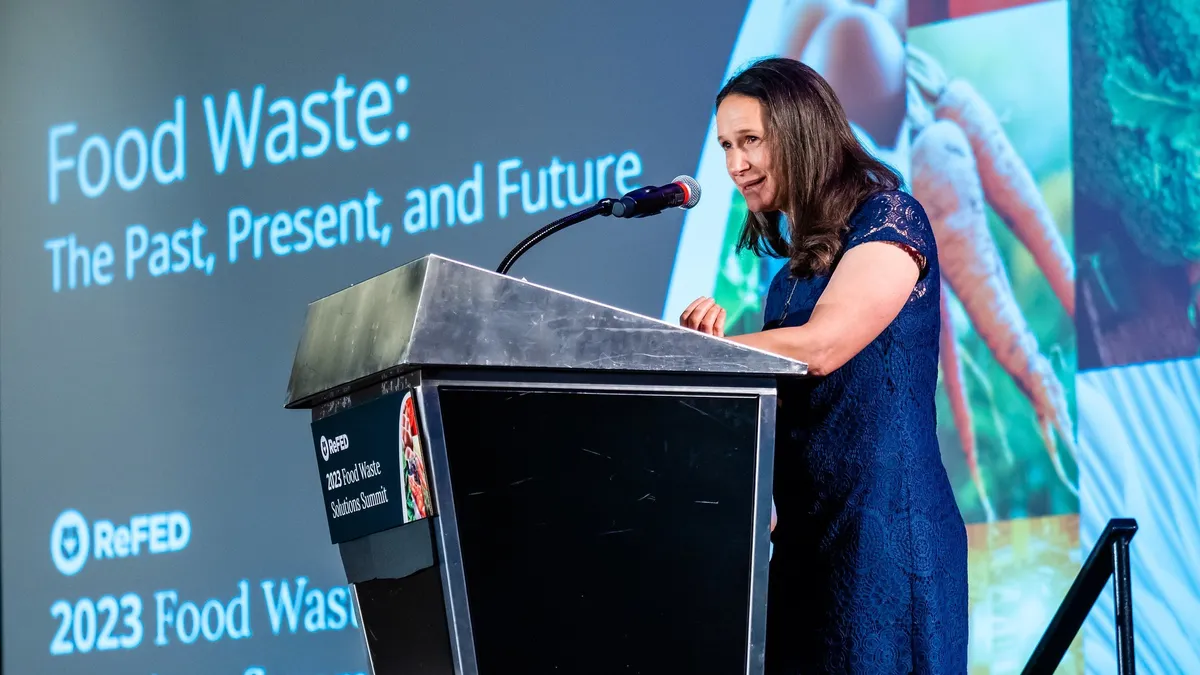 A woman speaks at a podium in front of a large projection screen that says, "Food Waste: The Past, Present and Future"