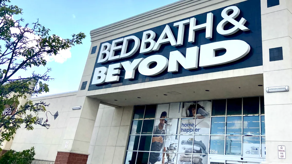 White clouds hover over a Bed Bath & Beyond store, a leafy tree in front on the left.