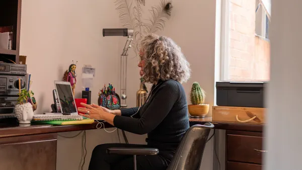 A person with curly gray hair sits a desk and works on a laptop.
