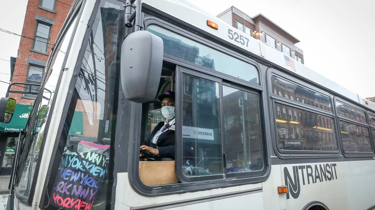 A bus driver wearing a mask looks toward the camera as the driver is driving a white NJ Transit bus.