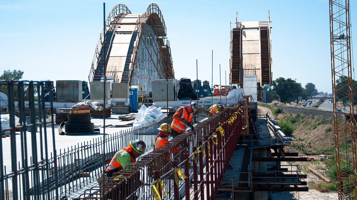 Four construction workers in reflective vests work on a partly-built concrete and metal bridge.