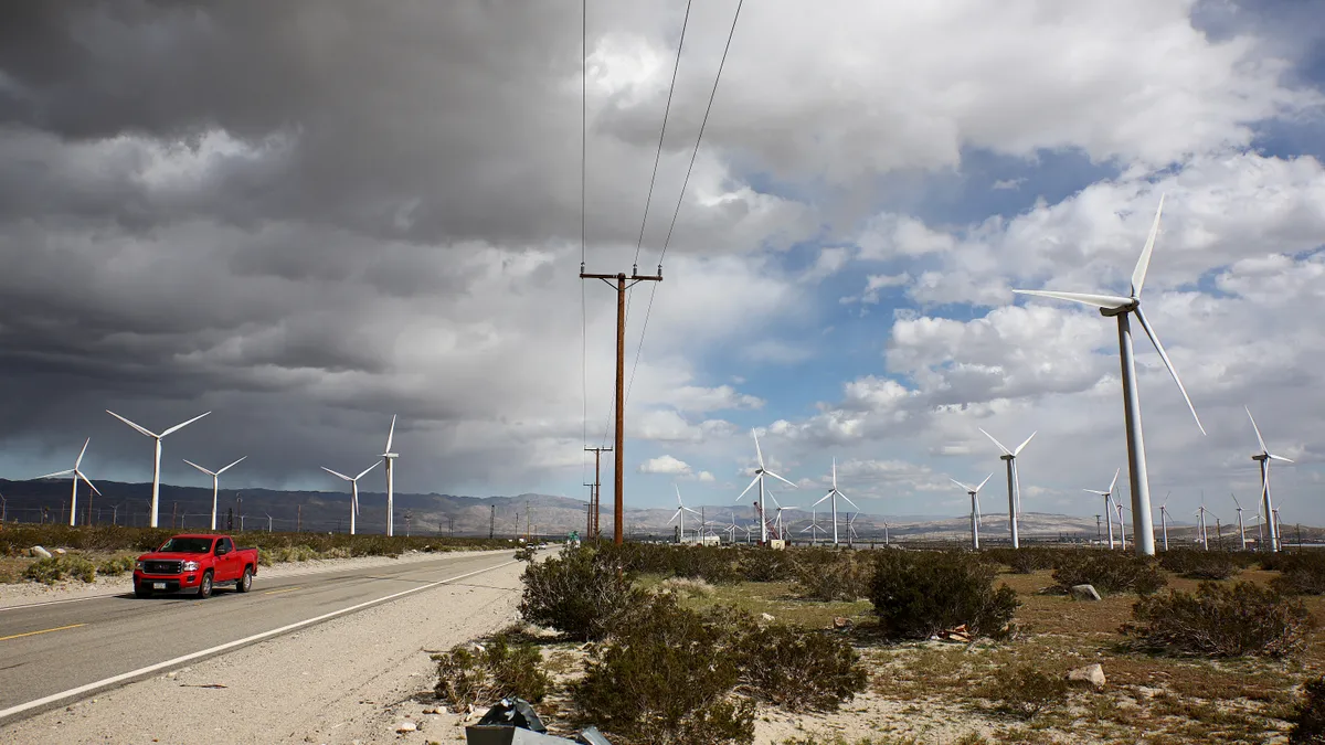 Truck driving along road with a field of wind turbines stretching across the land behind it and a storm coming in from the left side of the sky.