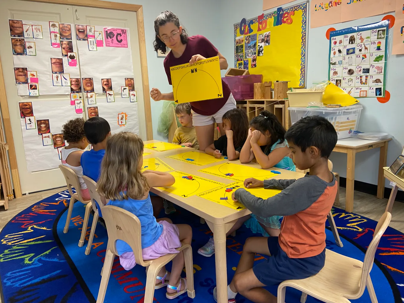 An adult stands next to a table of young students who are sitting in chairs. The adult has a book in their hands.