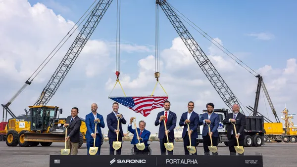 A group of people in suits pose with shovels above a box of dirt.