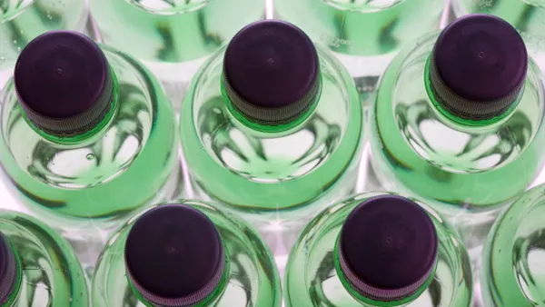 Overhead view of rows of green plastic sparkling water bottles with purple screw caps, arranged in a symmetrical grid pattern and illuminated from below, creating a vibrant, glowing effect.