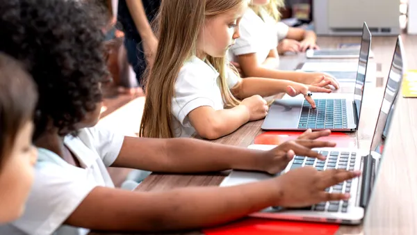 A handful of students are sitting at a table in a classroom. Each student has a laptop and is looking at the screens.