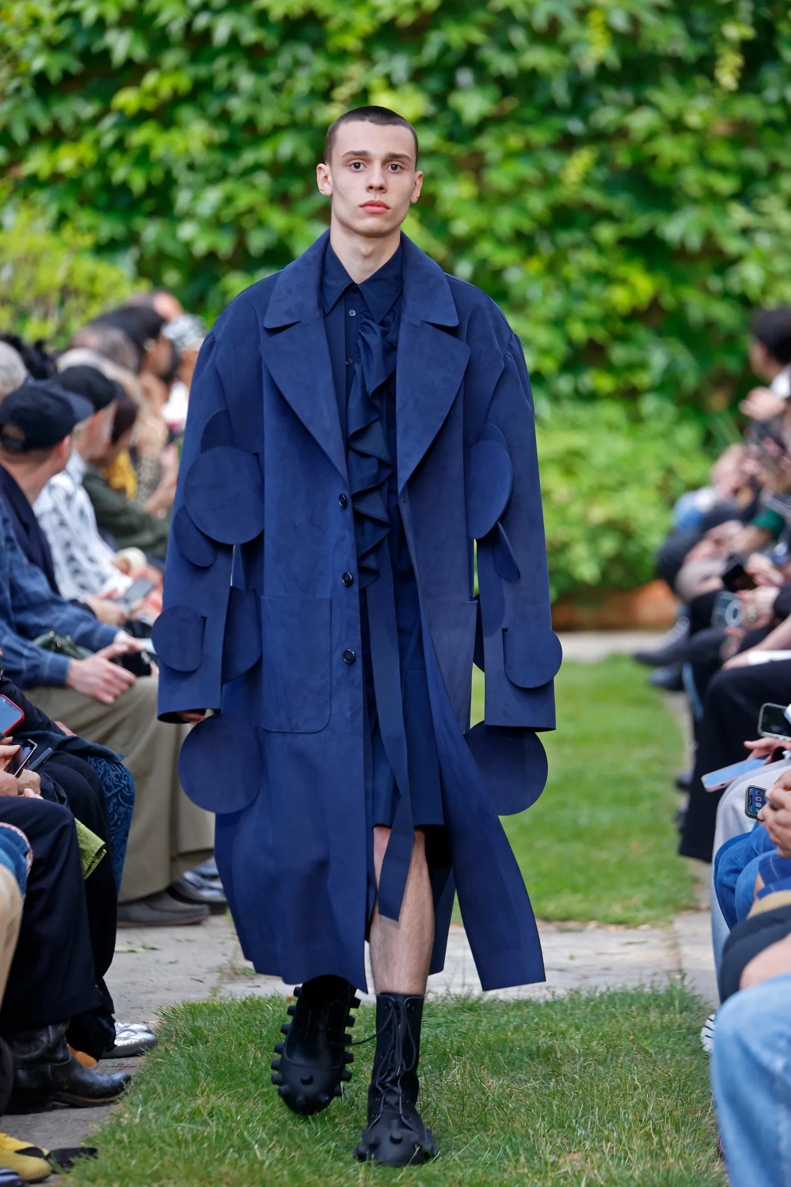 A model in a denim outfit covered in denim circles walks on a garden runway.