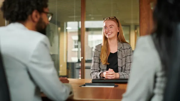 Three people are sitted at a desk. Two of them have their backs to the camera and one person is facing the camera and smiling with their hands on the desk.
