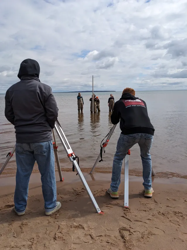Three students standing in Lake Superior with surveying equipment.
