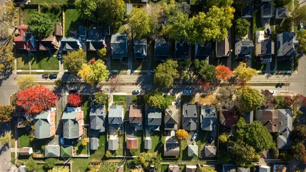 An aerial still of a residential neighborhood showing the roofs of single-family homes, lush trees and grass on a sunny day.