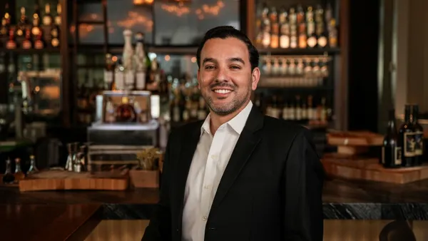 A smiling white man in a dark suit with no tie in front of a bar.