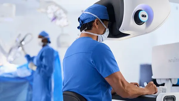 A surgeon sits at the console of a da Vinci 5 robot while another medical professional works in the background.