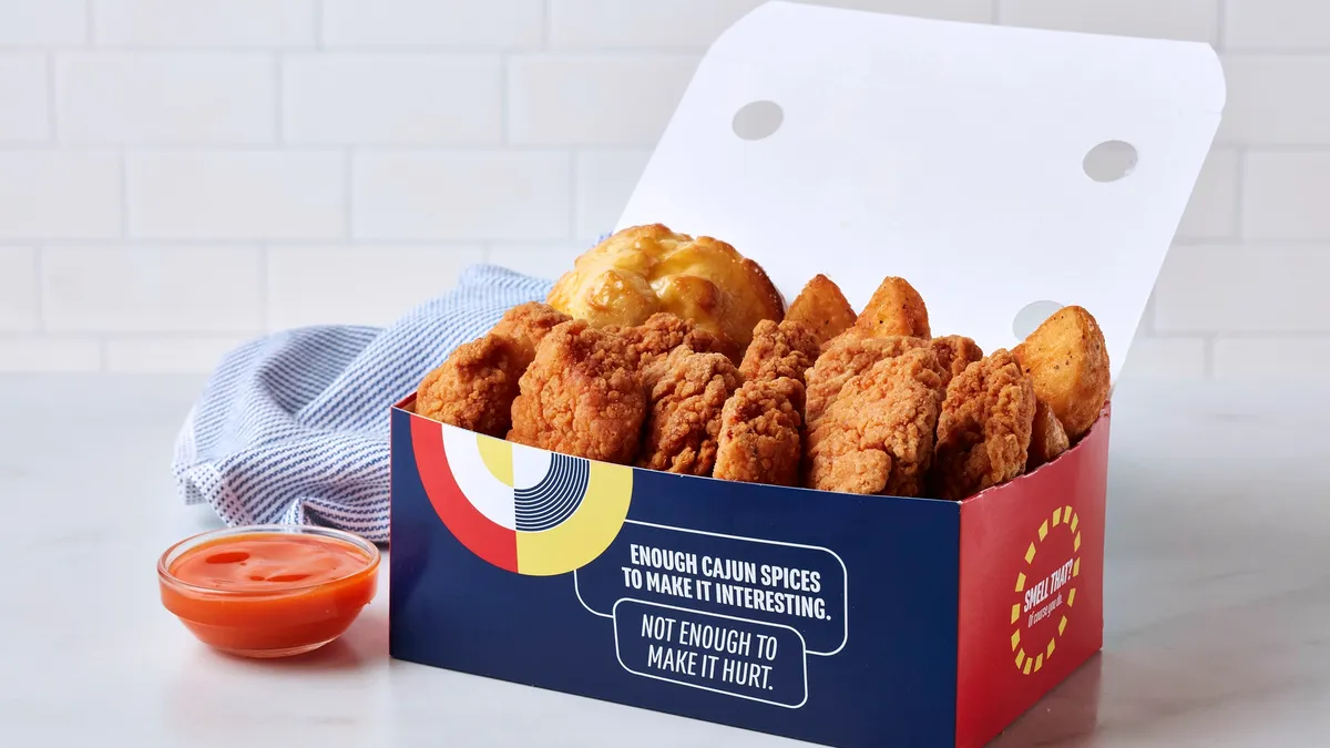 A photo of a box of chicken nuggets, potatoes and biscuits. On the front of the box, in white text on blue, it says "enough Cajun spices to make it interesting. Not enough to make it hurt." There is also a glass bowl of dipping sauce and a blue and white napkin on the white table next to the box.