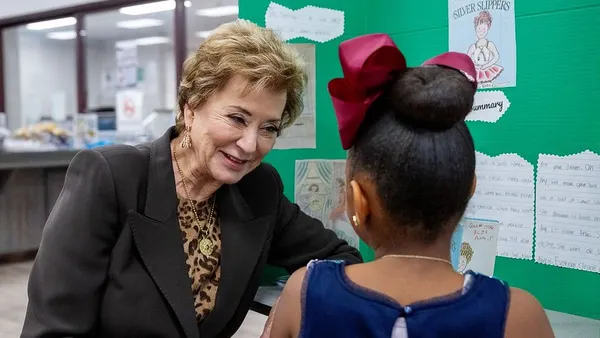 An adult sits at a table near a wall in a classroom. A student is sitting across from McMahon with their back to the camera