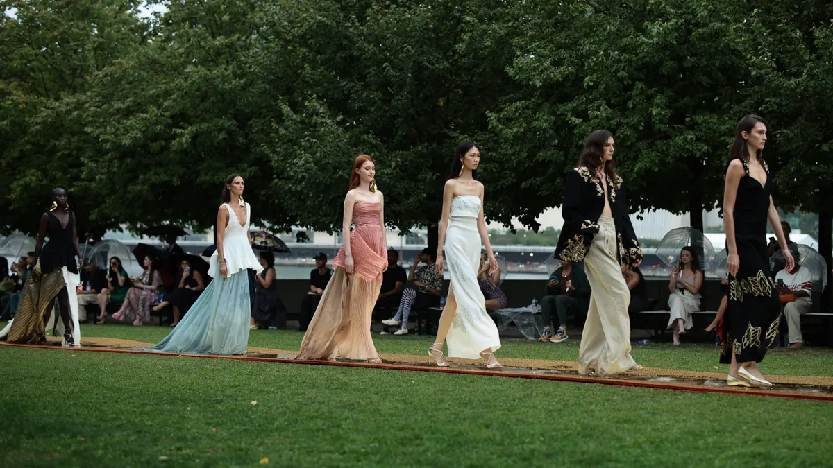 Models in long sheer dresses walk the outdoor runway at the Prabal Gurung fashion show during New York Fashion Week, September 2023, while the audience watches, holding transparent umbrellas.