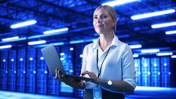 Person standing in a computer control room holding a laptop.