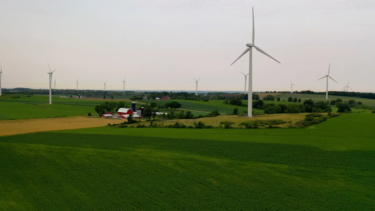 Wind turbines rise above a farm with green fields.
